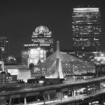 Black and white panoramic view of a city skyline at night with illuminated buildings and a curved, lit highway leading into the city.