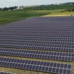 Aerial view of a large solar panel farm adjacent to agricultural fields under a cloudy sky.