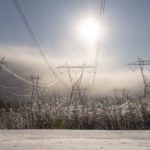 Sun shining above snow-covered power lines and electricity pylons in a wintry landscape.