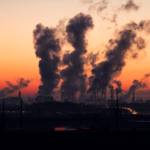 Industrial landscape at sunset, with silhouettes of multiple smokestacks emitting thick smoke against a vibrant orange sky.