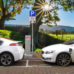 Two electric cars charging at a station with solar flare and green countryside background.