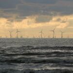 Silhouette of offshore wind turbines against a cloudy sunset sky, viewed from a wavy sea.