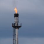 A gas flare burning brightly at the top of a metal industrial chimney against a cloudy sky.