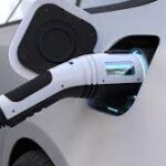 Electric vehicle charging cable connected to a white car, displaying the battery level on the plug.