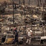 Firefighters walk through a devastated area with charred vehicles and remains of burnt structures, indicating a severe wildfire aftermath.