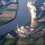 Aerial view of a nuclear power plant with smoke emitting from a cooling tower, surrounded by a river and agricultural fields.