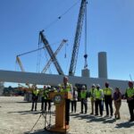 President speaking at a podium with a group of workers in hard hats at a construction site for wind turbines, with large crane and parts in the background.