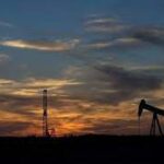 Silhouette of an oil pump jack against a colorful sunset sky with faint clouds.
