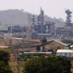 Industrial landscape showing several large factory structures with chimneys, surrounded by sparse vegetation and a backdrop of hills.