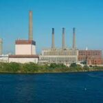 Large industrial power plant with multiple chimneys by a river under a clear blue sky.