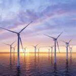 Offshore wind turbines stand in ocean waters against a sunset sky with soft pastel colors.