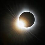 A solar eclipse with the moon partially covering the sun, creating a bright corona visible in a dark sky.