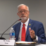 Elderly man with a beard, wearing glasses and a suit, speaking at a table with nameplate "mr. weber texas" and an american flag in the background.